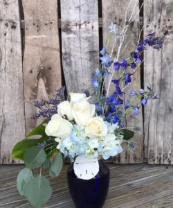 Towering blue delphinium accent this sapphire blue vase with a delicate sand dollar tied to the vase. Puffs of hydrangea and white roses make the focal point and the design is finished with silver dollar eucalyptus foliage.